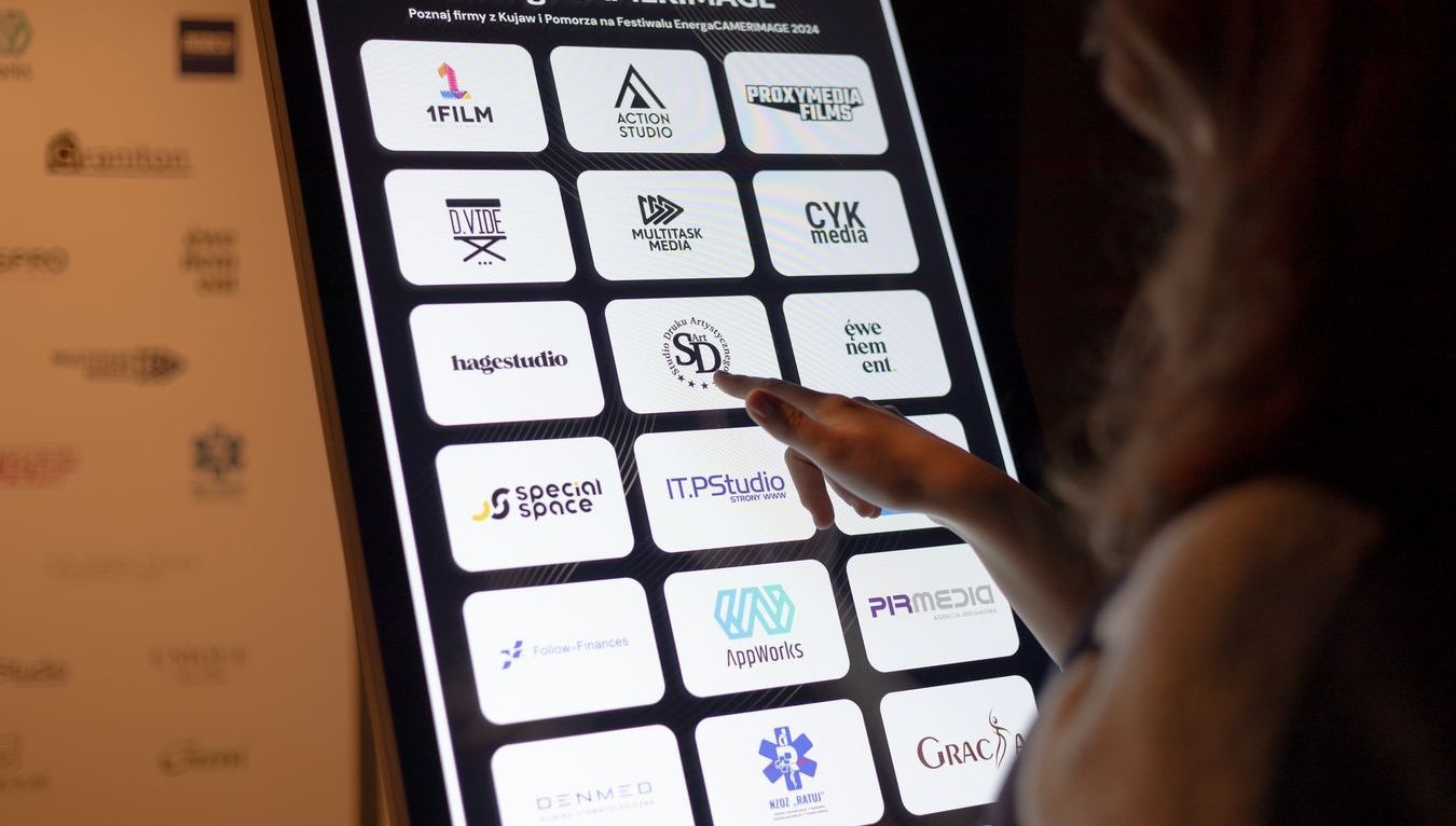 Osoba klika w logotyp znajdujący się na monitorze infokiosku. 